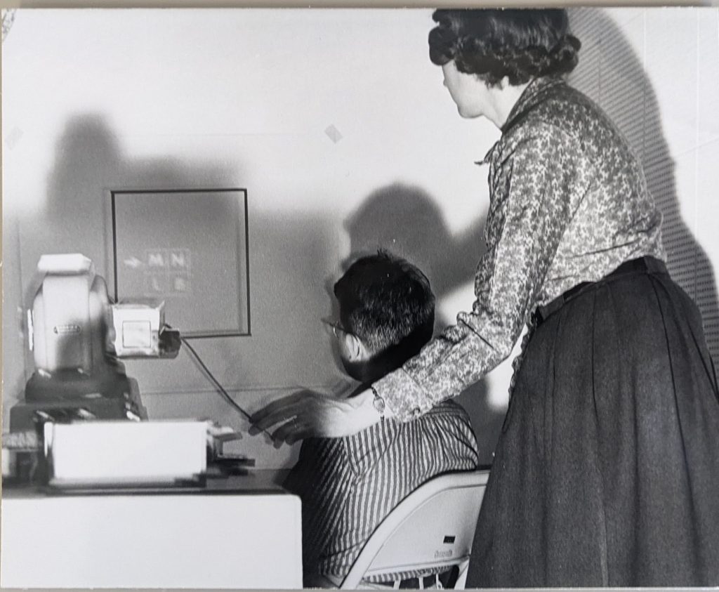 Young child seated in a room with a projector displaying a logic puzzle, while a researcher stands nearby controlling the projector