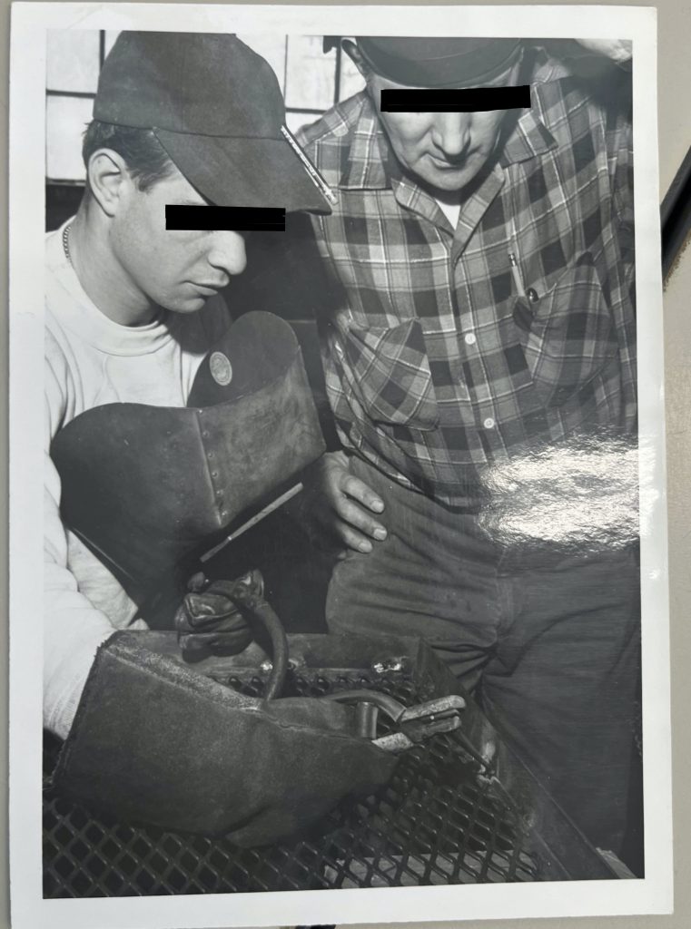 Experienced welder supervising younger man holding electrode holder while attaching wire screen to a frame