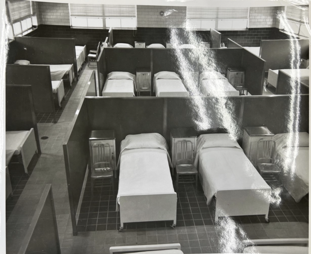 Dormitory with beds arranged closely, separated by chairs and short dividers